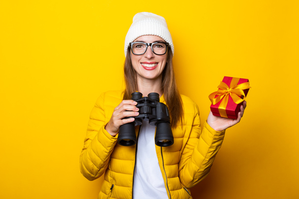 Smiling young woman in yellow jacket holding binoculars and gift on yellow background - Photo, Image