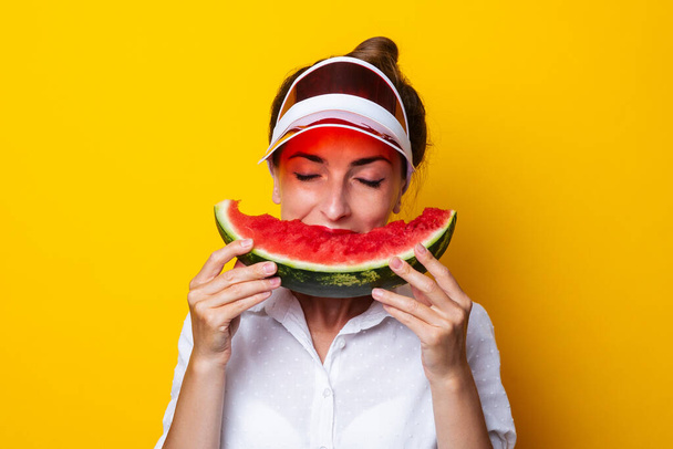 Young woman in red visor with closed eyes bites a slice of watermelon on yellow background - Photo, image