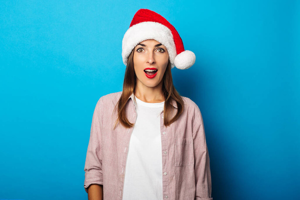 Überraschte junge Frau im Hemd mit Weihnachtsmann-Hut auf blauem Hintergrund - Foto, Bild
