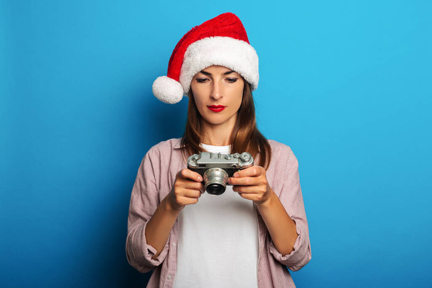 Young woman in santa claus hat looks thoughtfully at a retro camera on a blue background - Fotoğraf, Görsel