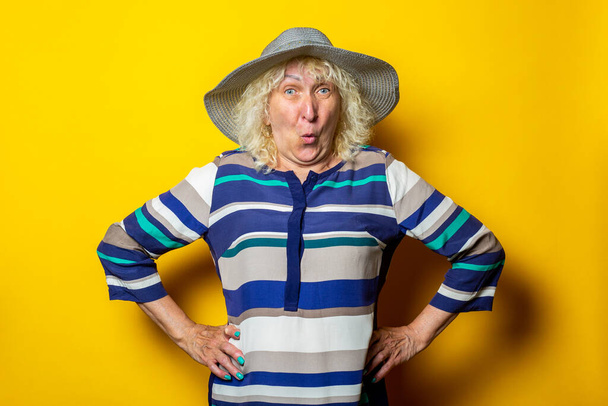 Überraschte alte Frau in Kleid und Hut hält ihre Hände an der Taille auf gelbem Hintergrund. - Foto, Bild