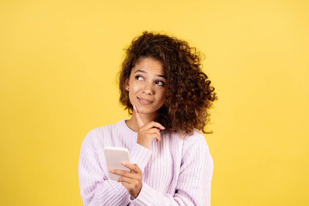 Portret van een bedachtzame jonge Afrikaans-Amerikaanse vrouw die opzij kijkt, twijfelt, een moderne smartphone in handen houdt en op een gele kopieerruimte staat - Foto, afbeelding