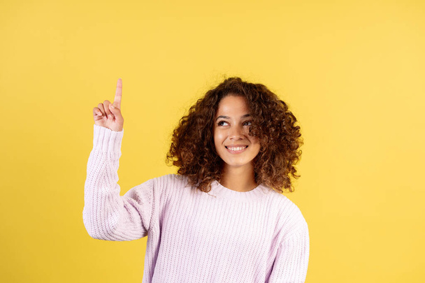 Happy student girl has an idea. Smiling adult afro american woman pointing up with finger, showing solution and standing on yellow copy space background - Фото, зображення