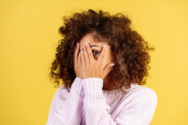 Portrait of scared or curious adult afro american woman cover face with hands, peeks through the palm, looking at camera and standing on yellow copy space background - Photo, Image