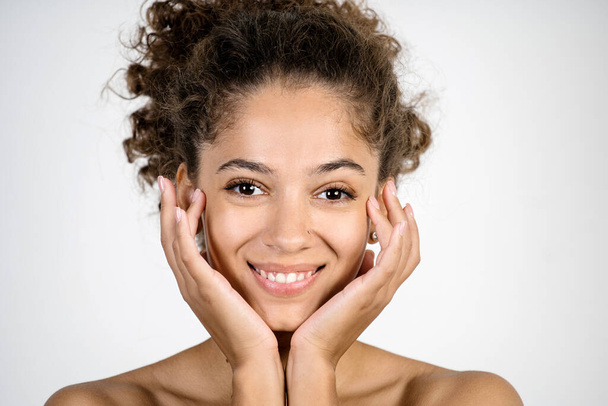 Concetto di cura della pelle. Ritratto facciale di felice giovane donna afro-americana che si tiene per mano vicino alla pelle pulita e fresca, guardando la fotocamera, sorridente ampio, in piedi isolato su sfondo bianco con spazio copia - Foto, immagini