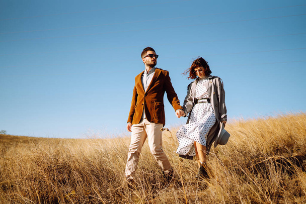 Stylish  young couple enjoying autumn weather in autumn field. Lovely couple walking and hugging in the field together. The concept of youth, love and lifestyle. - Фото, изображение