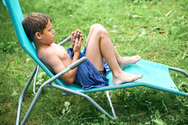 Adicción a los artilugios. Niño mojado sentado en la tumbona y jugando en el teléfono inteligente después de nadar en la piscina al aire libre - Foto, Imagen