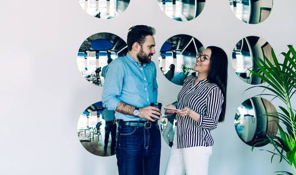 Positive smiling coworkers in trendy casual wear standing in contemporary light office while relishing warm beverage and using smartphone and communicating - Φωτογραφία, εικόνα