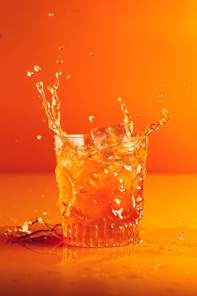 IJs valt in een geslepen kristalglas met oranje vloeistof en veroorzaakt een plons op een oranje achtergrond - Foto, afbeelding