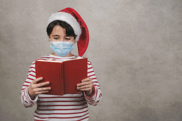 Merry Christmas,kid with medical mask Wearing Christmas Santa Claus hat reading a book over gray background - Фото, изображение