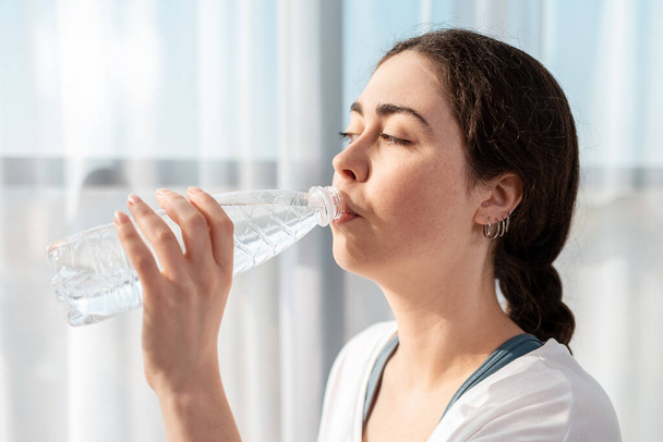 Portret van een jonge vrouw die een fles water drinkt. Grote ramen op de achtergrond. Gezonde levensstijl. - Foto, afbeelding