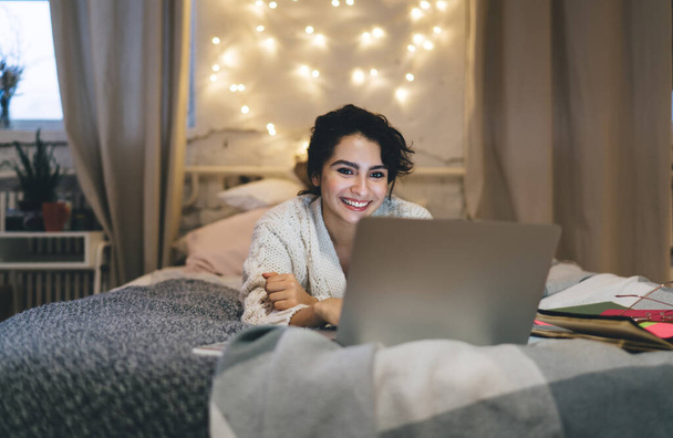 Optimistic female freelancer smiling and typing on laptop keyboard while working on remote project and lying on comfortable bed at home - Foto, Imagen