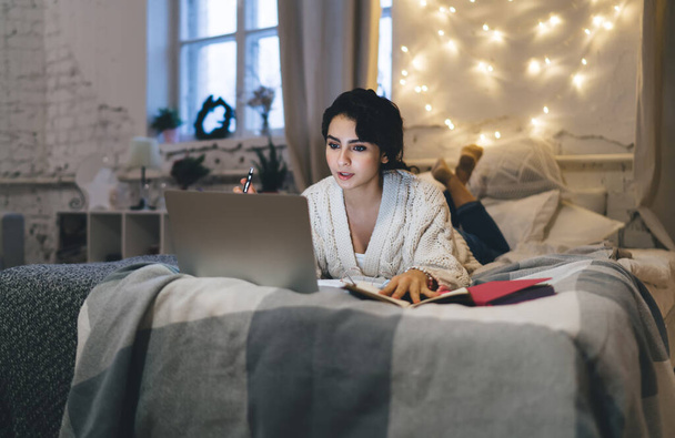 Full body pensive woman in pajama lying on comfortable bed and working on remote project on laptop at home during weekend - Фото, изображение