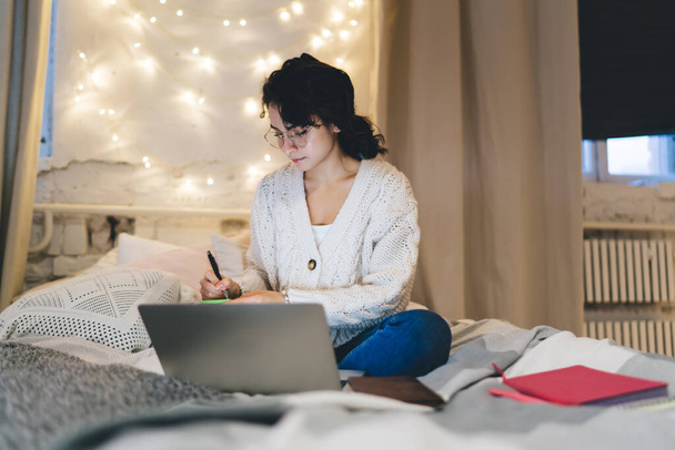 Сосредоточенная женщина-фрилансер в свитере и очках сидит на удобной кровати и делает заметки в блокноте во время работы на ноутбуке - Фото, изображение