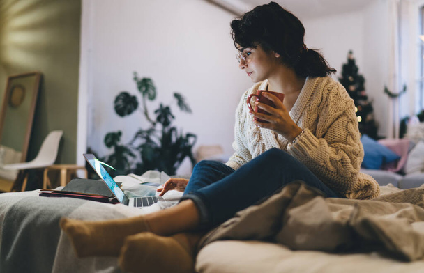 Low angle of focused woman in eyeglasses and casual clothes sitting on comfortable bed and drinking cup of coffee during work on laptop - Foto, imagen