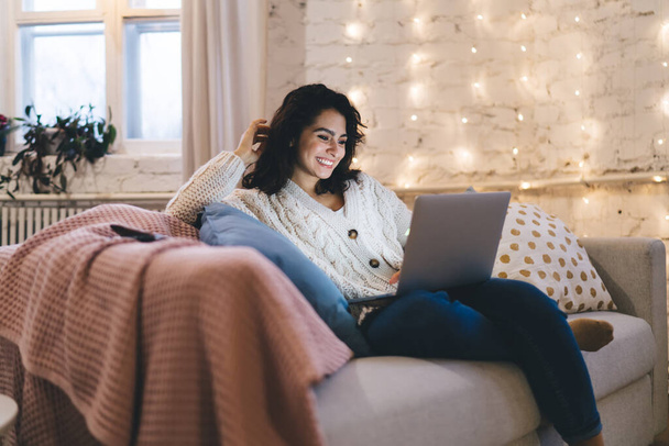 Positive attractive female in warm clothes sitting on comfortable sofa in living room at home and smiling while browsing laptop - Foto, afbeelding