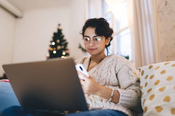 From below of content young female in warm clothes and eyewear sitting on sofa with laptop and mobile phone in Christmas Day - Zdjęcie, obraz
