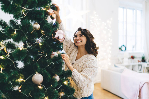 Delight young lady standing near decorated with baubles and garlands Christmas tree on cozy room with star and lights on wall and window - 写真・画像