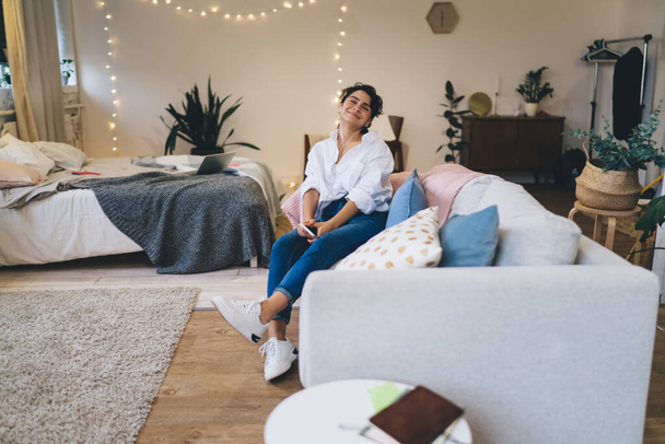Full body cheerful female in casual outfit smiling and looking at camera while resting on comfortable sofa in living room - Фото, изображение