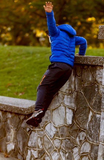 A hyperactive kid is jumping off a high wall as he is running away. The courageous boy wears track pants, sneakers and a hooded coat. He is full of energy. He extends his arm for balance. - Foto, Imagen