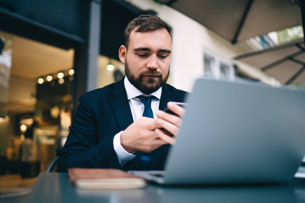 Concentrated bearded businessman wearing formal clothes looking at smartphone while working on business project in office with laptop and notebook - Fotoğraf, Görsel