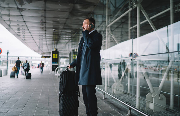 Caucasian businessman waiting near airport terminal using mobile phone for making international conversation with corporate partner, formally dressed boss communicate via mobile during work travel - 写真・画像