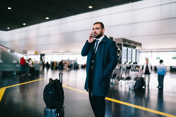 Handsome male lawyer with luggage suitcase using cellular gadget for communicate during business trip at airport, travelling businessman making phone call for discussing arrival corporate meeting - Fotó, kép
