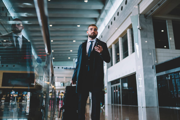 Kontemplacyjny dyrektor korporacyjny z bagażem walizkowym i technologią smartfonów przemyślane odwracając wzrok podczas wyszukiwania bramy terminala na lotnisku, pracodawca kaukaski 30 podczas podróży służbowej - Zdjęcie, obraz