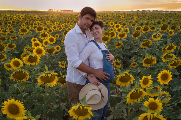 Fröhliches kaukasisches Paar beim Mutterschaftsshooting in einem Sonnenblumenfeld bei Sonnenuntergang - Foto, Bild