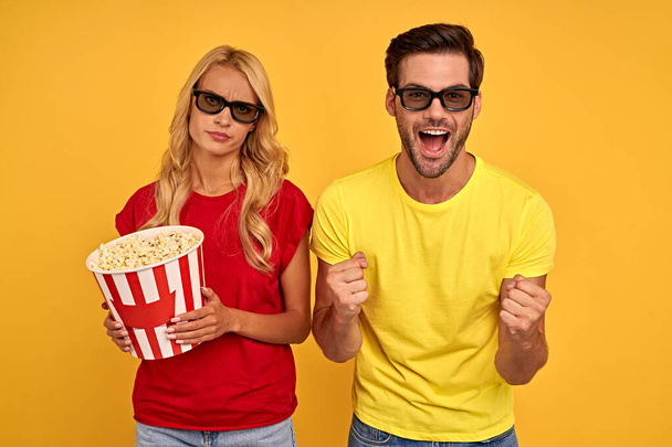 Joyful young couple two friends guy girl in 3d glasses isolated on yellow background. People in cinema lifestyle concept. Watching movie film, hold buckets of popcorn. - Фото, изображение
