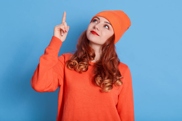 Winsome girl in hat and sweater pointing up at copy space and looking up with smile, adorable lady with wavy hair, standing isolated over blue background. - Photo, Image