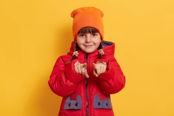 Close up of little dark haired kid with two pigtails, girl wearing red jacket and orange cap isolated on yellow background, pointing index finger to camera. - Foto, afbeelding