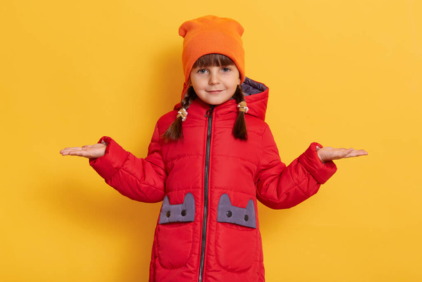 Female child spreading palms aside and looking directly at camera, posing isolated over yellow background, having two pigtails, wearing casual jacket and cap, copy apace for advertisement. - Photo, image