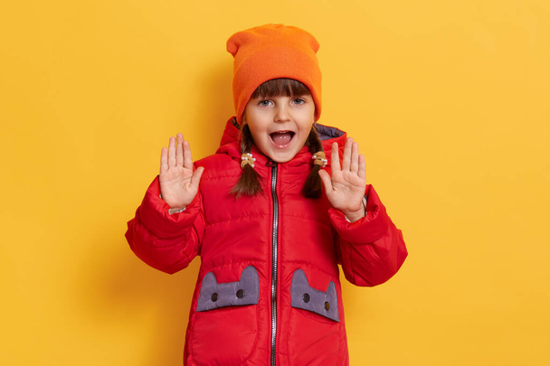 Little adorable girl wearing casual red jacket and orange cap, keeps mouth opened, yelling something, raising palms and doing stop gesture against yellow wall. - Fotografie, Obrázek