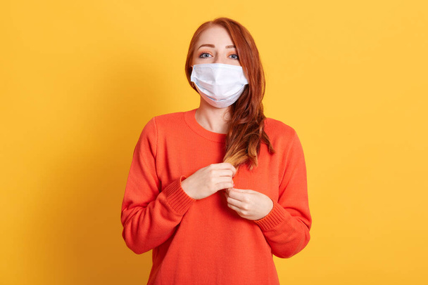Gorgeous red haired young female wearing protective mask against COVID virus, tired of stress, looks confidently at camera with fear in eyes, poses against yellow background. - Photo, Image