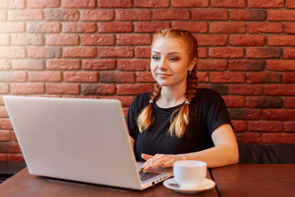 Business woman sitting against brick wall in coffee shop, working on her lap top, looking at device screen, enjoying hot coffee from white ceramic cup, student with red hair and two pigtails. - Photo, image