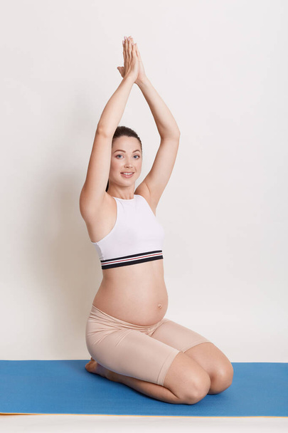 Portrait of pregnant woman doing yoga while sitting with raising palms on blue yoga mat, expectant mother looking smiling at camera, showing her bare belly. - Фото, изображение