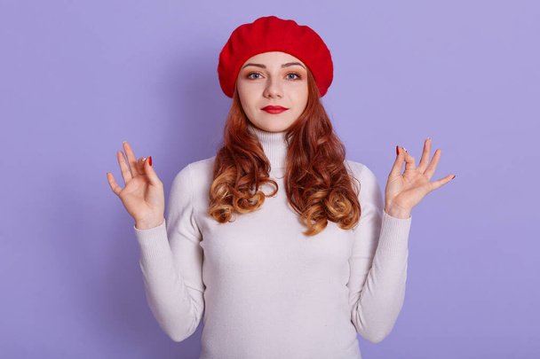 Image of young Caucasian girl wearing beret and white casual shirt showing okay sign, meditating, trying to relax, looks at camera wit calm facial expression. - Zdjęcie, obraz