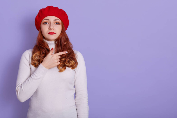Image of young displeased woman pointing finger aside and looking directly at camera with sad facial expression, copy space for promo, standing against lilac background. - Foto, imagen