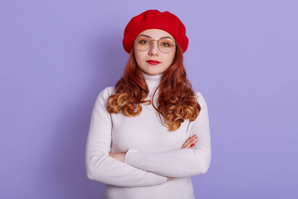 Portrait of lovely woman wearing white shirt and red beret, looking at camera with serious facial expression, stands with folded hands, looking strict at somebody. - Zdjęcie, obraz