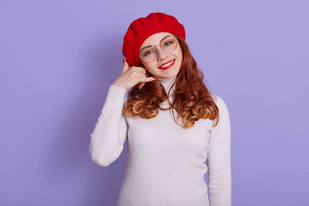 Woman in glasses doing phone gesture like says: call me back with hand and fingers like talking on telephone isolated on lilac background, adorable girl wearing red beret and white shirt. - Фото, зображення