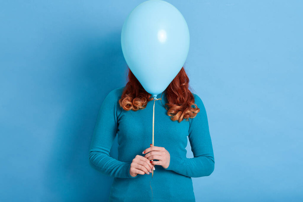 Portrait of woman hides her face behind helium balloon, red haired female with curls holding ballon in front her face against blue background, holiday, celebration. - Foto, Bild