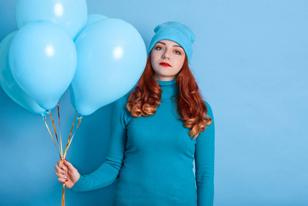 Sad and bored young European woman holding bunch of helium balloons while standing against blue background, woman with upset facial expression celebrating her birthday. - Фото, зображення
