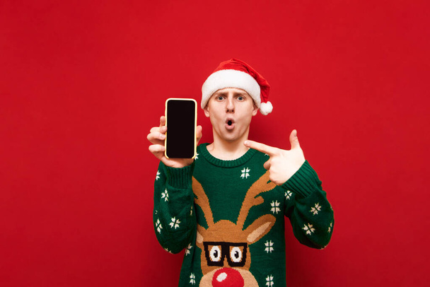 Shocked guy in santa hat and green sweater shows his finger on smartphone screen and looks into camera with surprised face on red background. Christmas and New Year concept. Copy space - Photo, Image