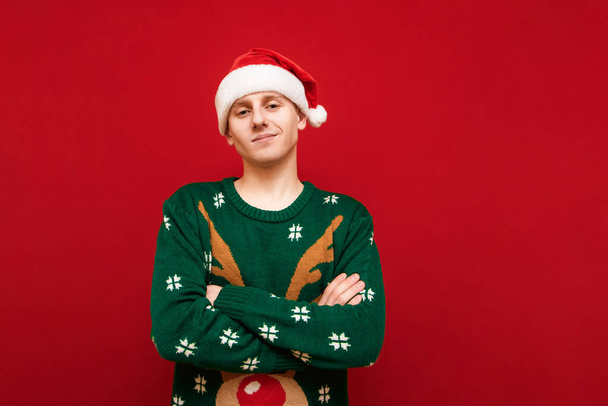 Leuke jongeman in kerstmuts en groene trui geïsoleerd op rode achtergrond, kijkend in de camera en glimlachend. Glimlachende man met kerst stemming geïsoleerd op rode achtergrond. - Foto, afbeelding