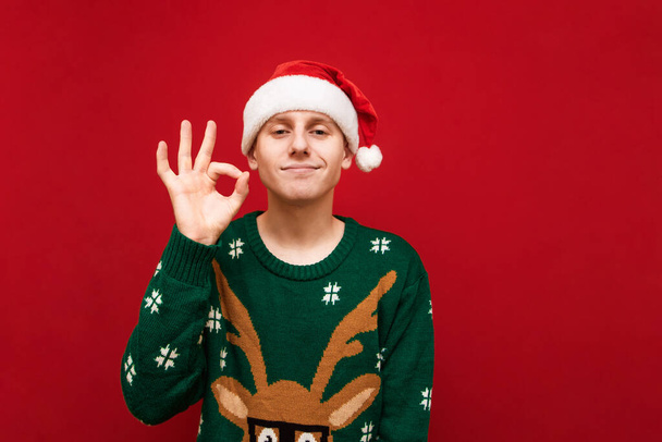 Funny young man in christmas clothes looks into the camera and shows ok gesture with smile on our face on red background. Positive guy in santa hat shows okay gesture, isolated. - Photo, Image