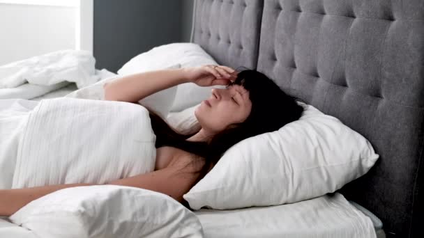 Jonge blanke vrouw kan niet slapen met hoofdpijn. Harde migraine veroorzaakt slapeloosheid. Meisje ligt verdrietig en slapeloos in de slaapkamer en worstelt op witte lakens, raakt het hoofd en zucht. - Video