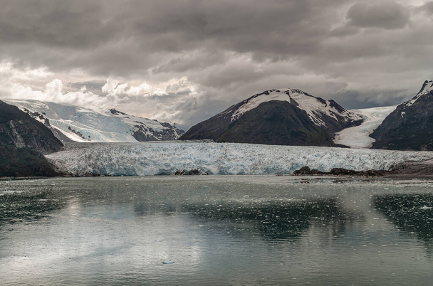 Sarmiento Channel, Chile - December 11, 2008: Long shot on Amalia Glacier ending in gray water and sliding between black rocky mountains under stormy brown cloudscape. Floating ice pieces. - Photo, Image