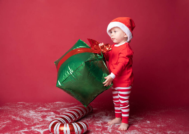 Bambino ragazzo in rosso cappello di Babbo Natale con bastoncino di zucchero natalizio e regali su uno sfondo rosso. - Foto, immagini