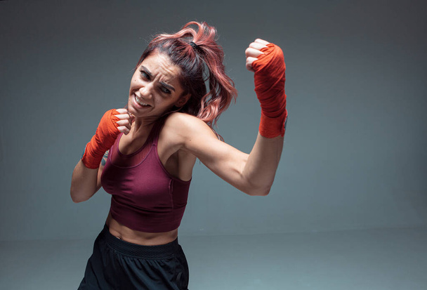 Pretty girl fighter in boxing bandages makes an uppercut in studio isolated on gray background. Strength and motivation - 写真・画像